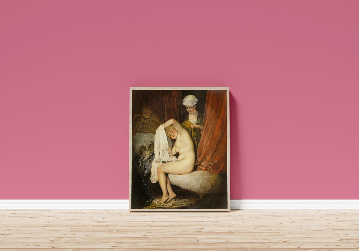 Framed art print of A Woman at Her Toilet by Jean-Antoine Watteau, in a light wood frame leaning on a pink wall above a wooden floor