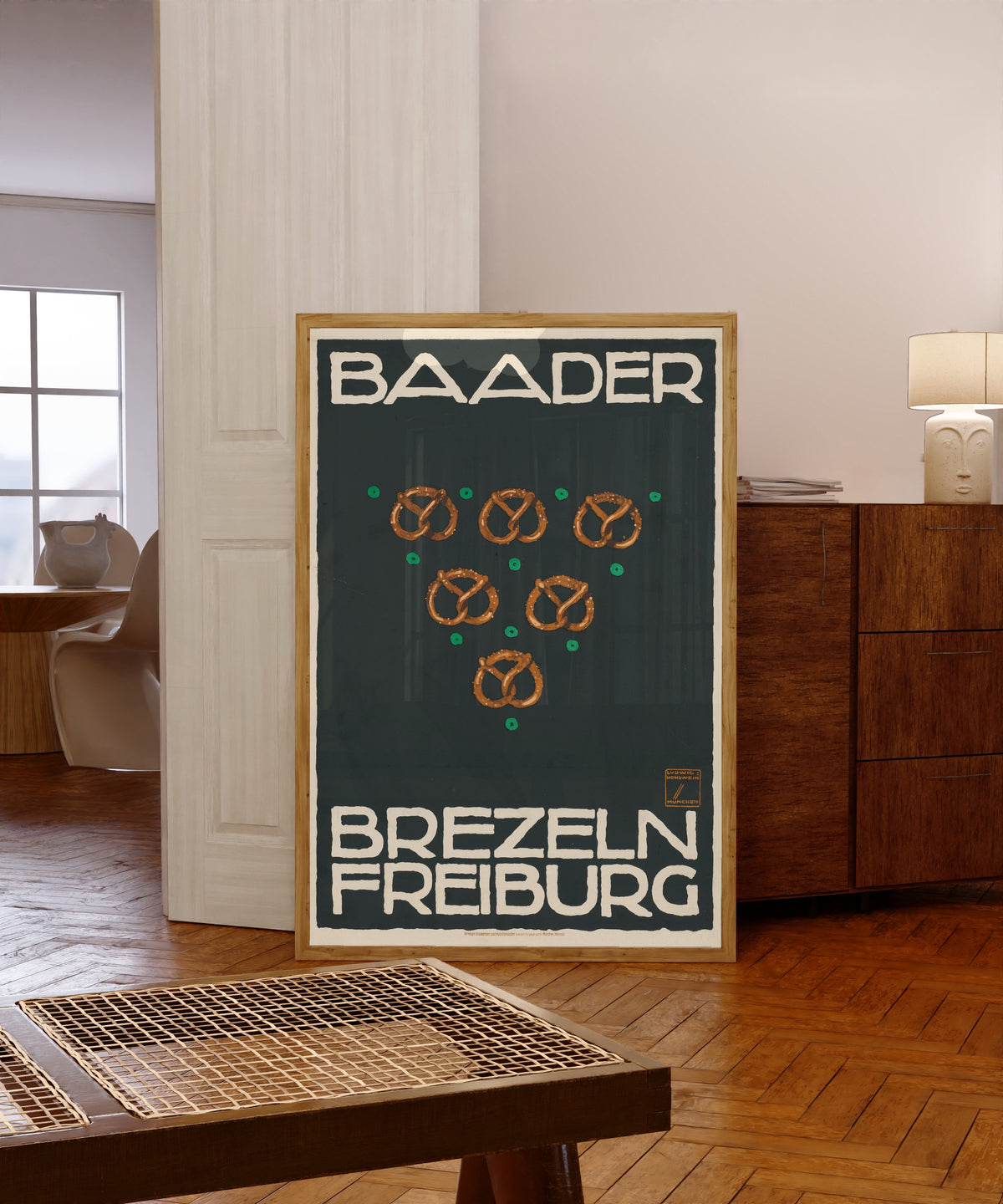 Framed art print of Baader Brezeln, Freiburg by Ludwig Hohlwein, in a light wood frame leaning in a warm room with a parquet floor