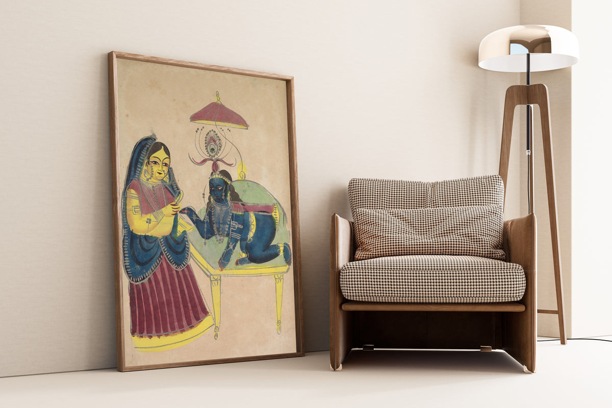 Framed art print of Baby Krishna Asking for Butter from Yashoda by Kalighat painter, leaning against a wall beside an armchair and a floor lamp