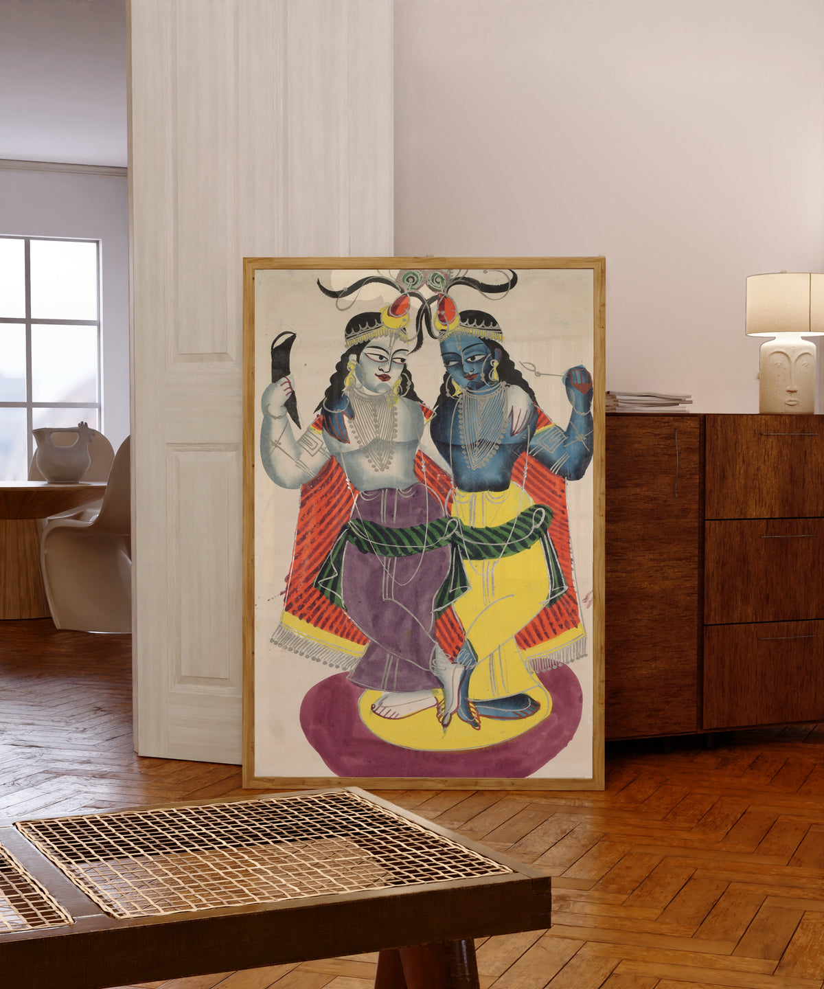 Framed art print of Balarama and Krishna by Kalighat painter, in a light wood frame leaning in a warm room with a parquet floor
