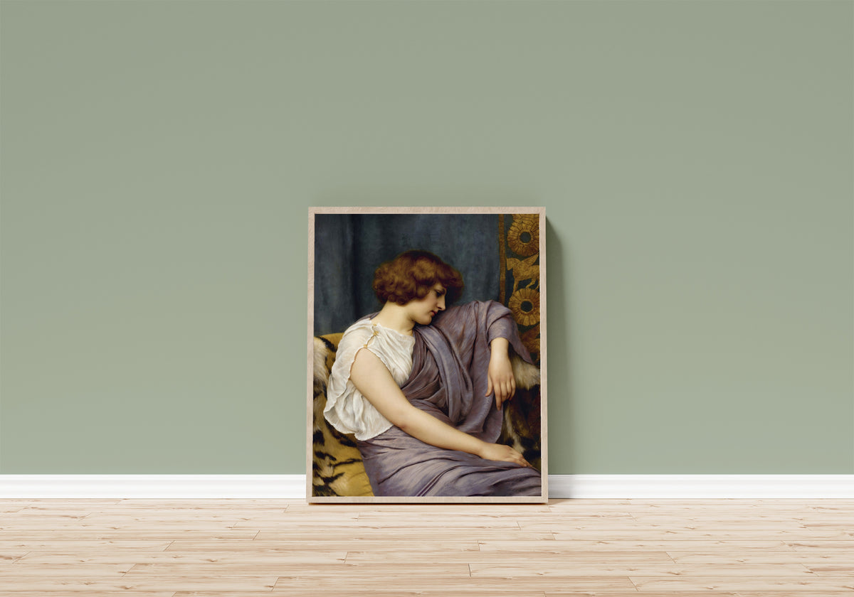 Framed art print of Briseis by John William Godward, in a light wood frame leaning on a sage green wall above a wooden floor