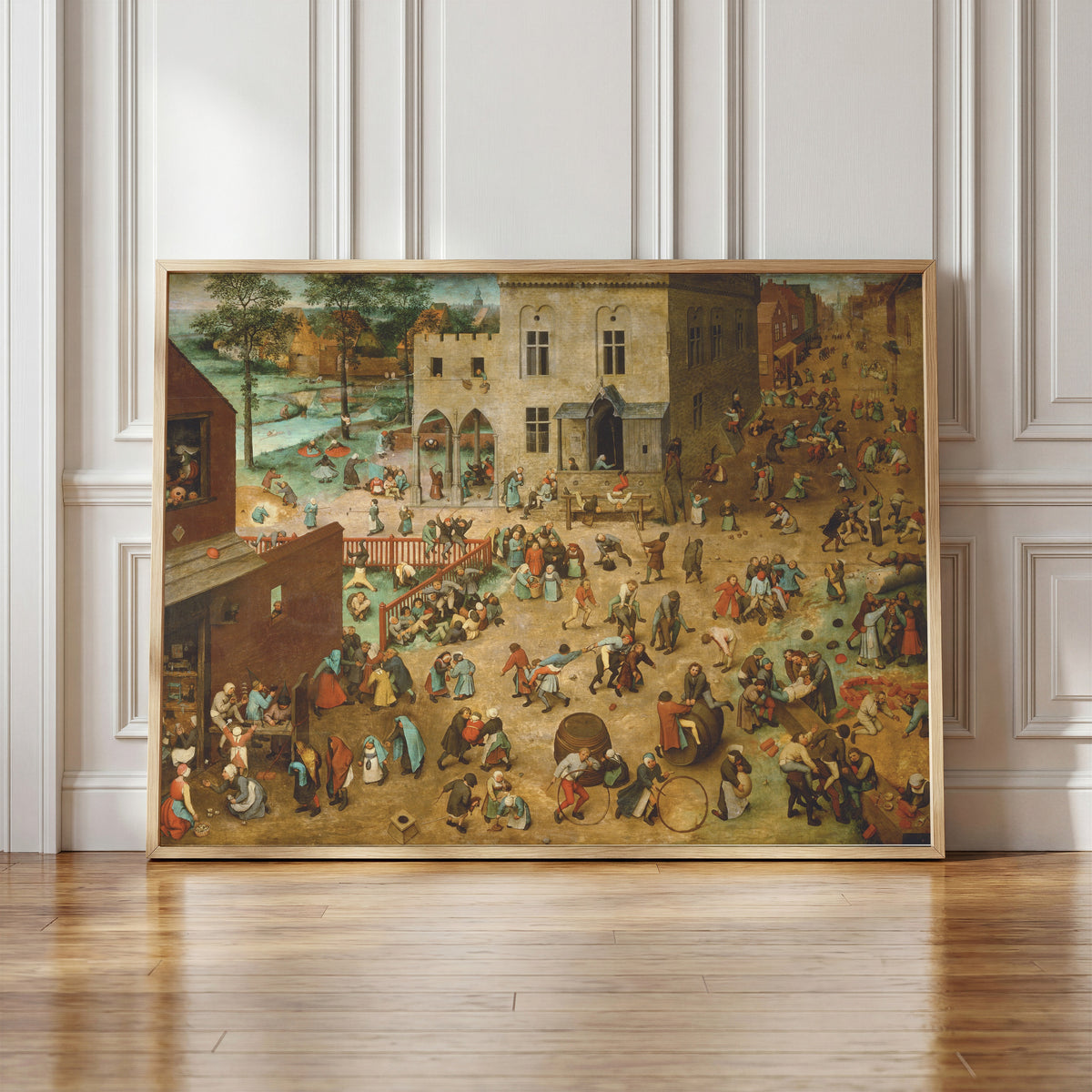 Framed art print of Children's Games by Pieter Bruegel the Elder, in a light wood frame leaning against a panelled white wall