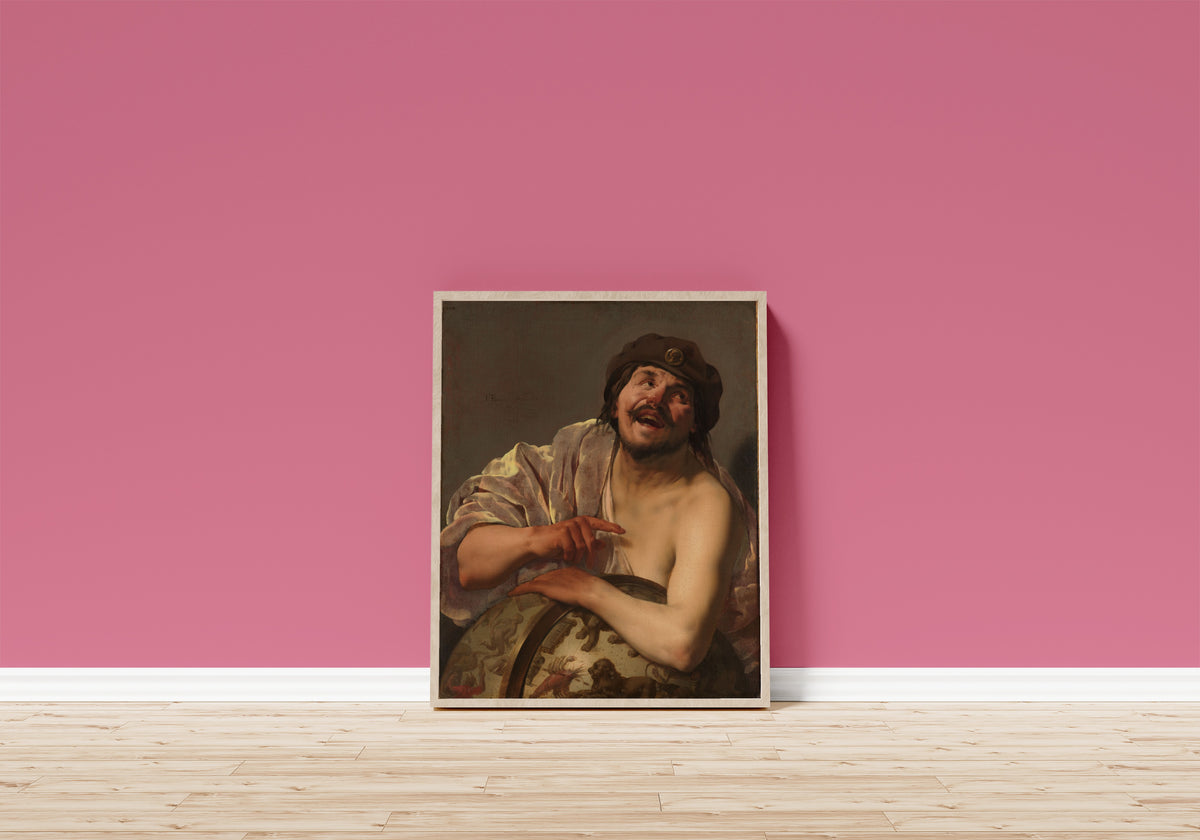 Framed art print of Democritus by Hendrick ter Brugghen, in a light wood frame leaning on a pink wall above a wooden floor