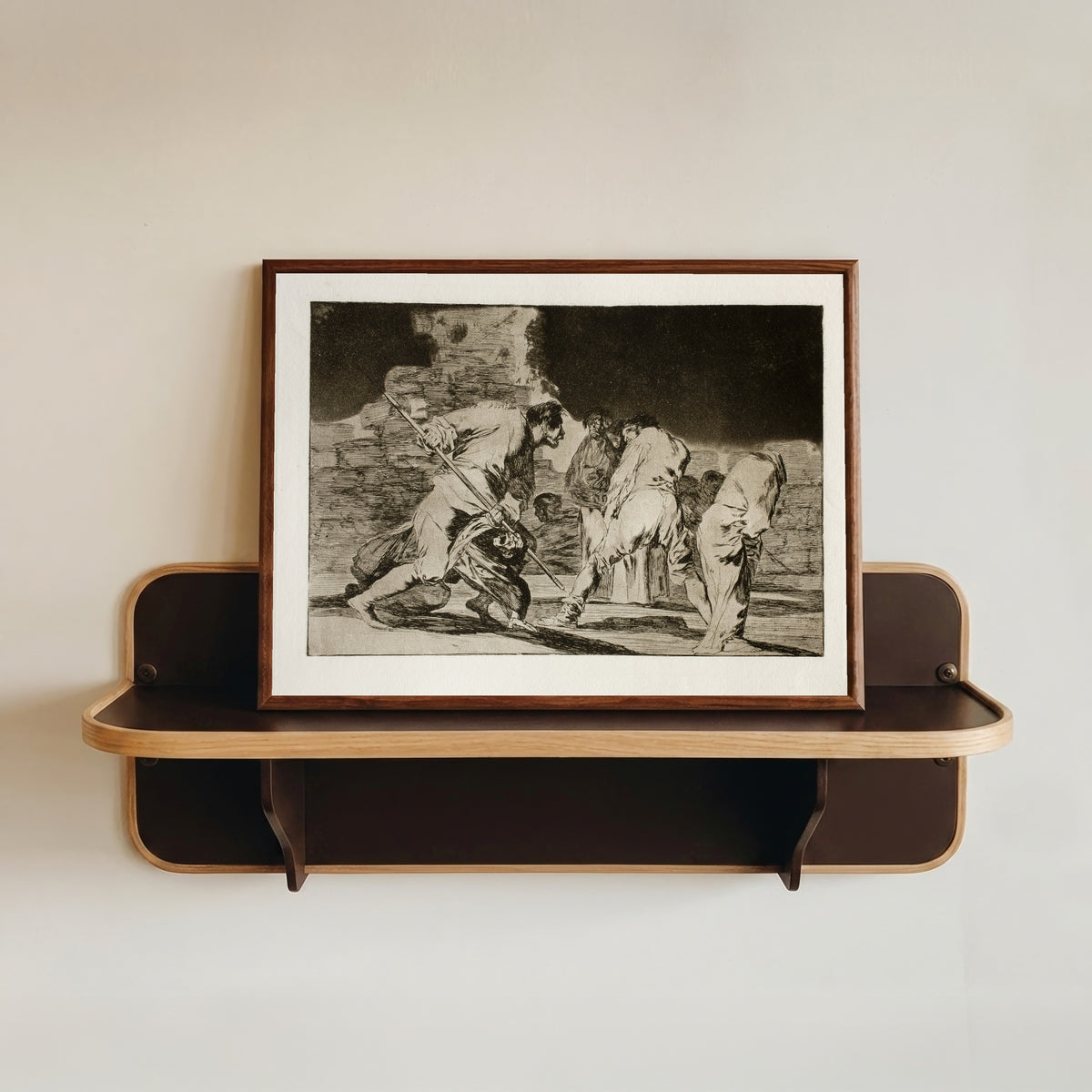 Framed art print of Disparate cruel (Cruel Folly), from Los Disparates by Francisco de Goya, in a dark wood frame resting on a wall shelf