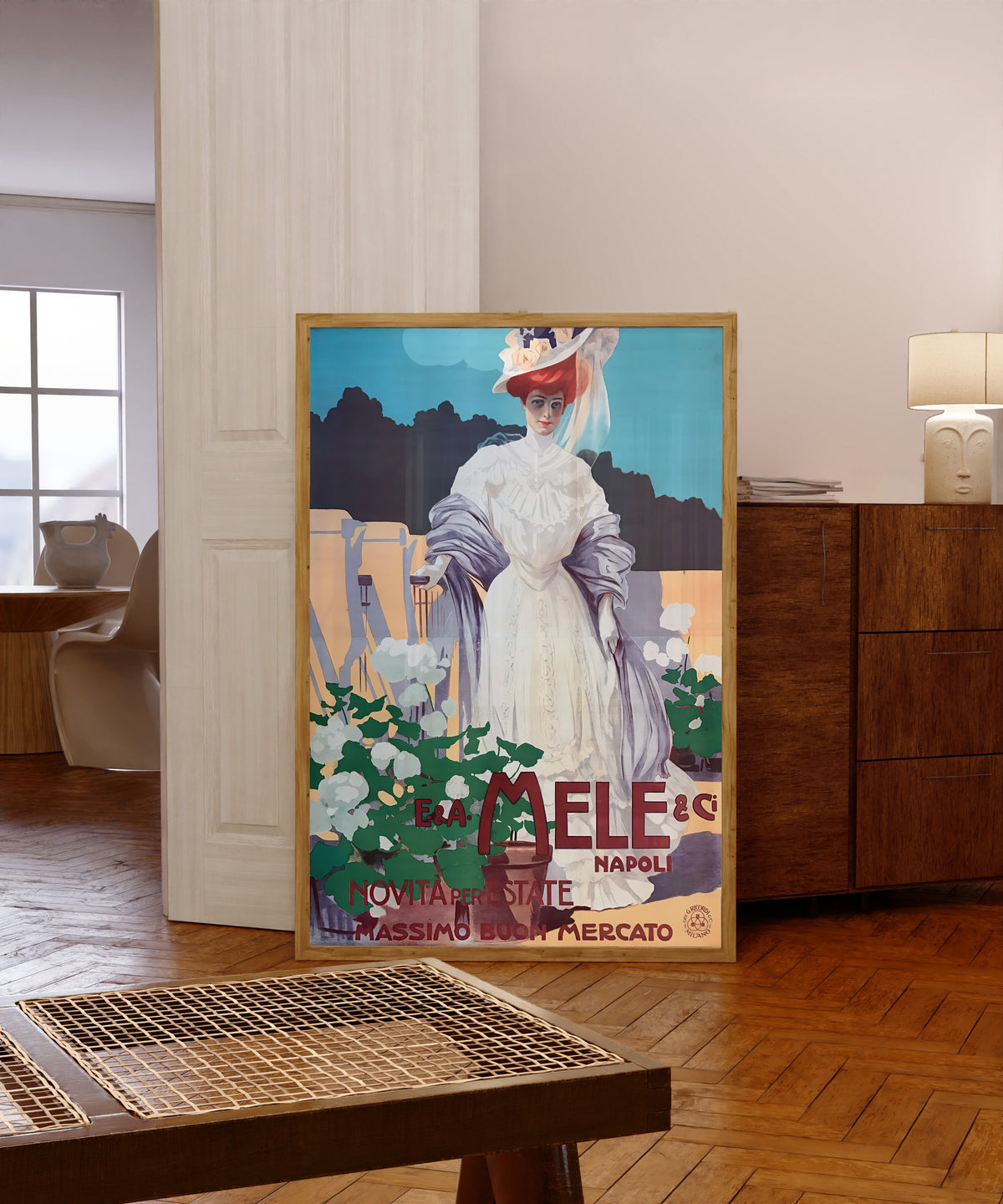 Framed art print of E. & A. Mele, Napoli: Novità per l'Estate… by Leopoldo Metlicovitz, in a light wood frame leaning in a warm room with a parquet floor