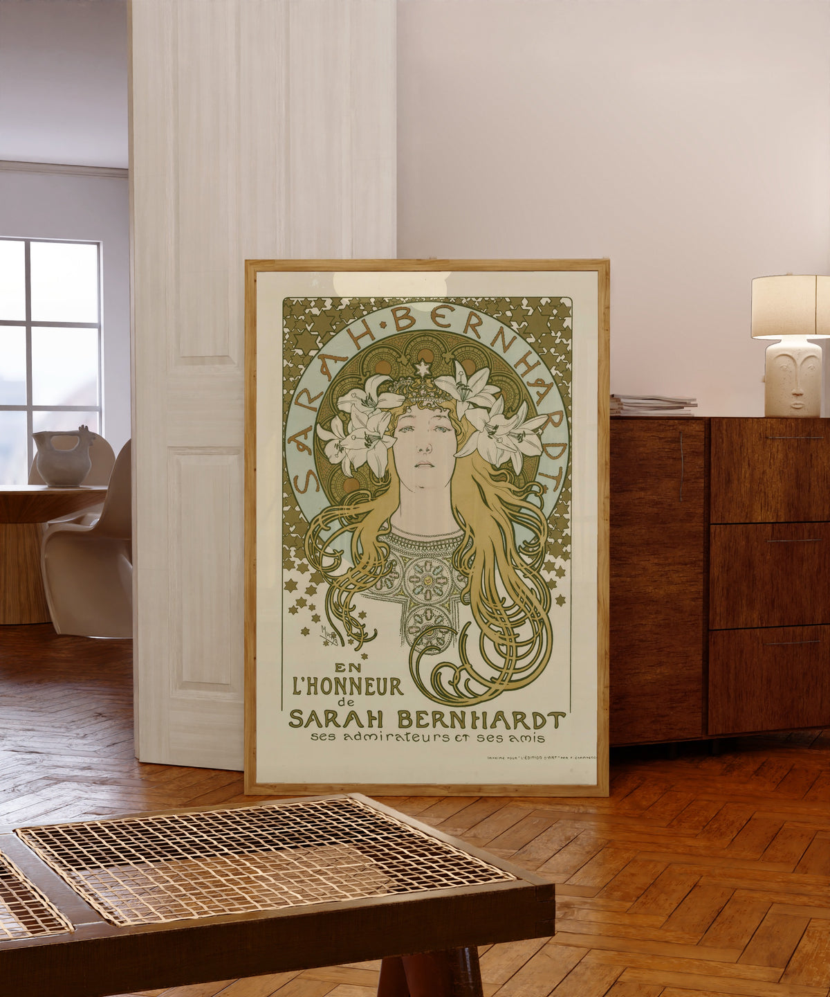 Framed art print of En l’honneur de Sarah Bernhardt, ses admirateurs et… by Alphonse Mucha, in a light wood frame leaning in a warm room with a parquet floor