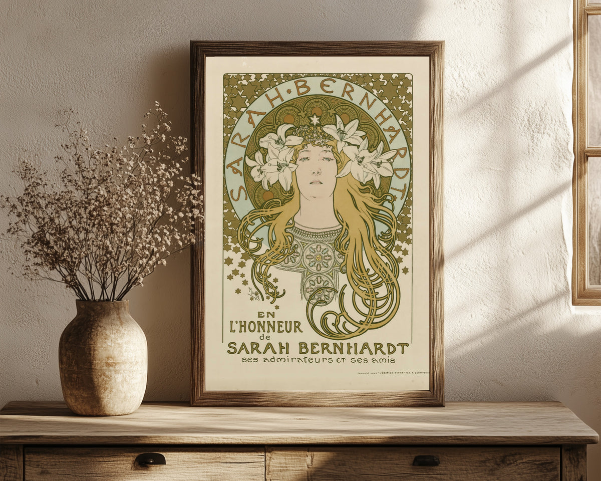 Framed art print of En l’honneur de Sarah Bernhardt, ses admirateurs… by Alphonse Mucha, in a dark wood frame leaning on a rustic console beside dried flowers