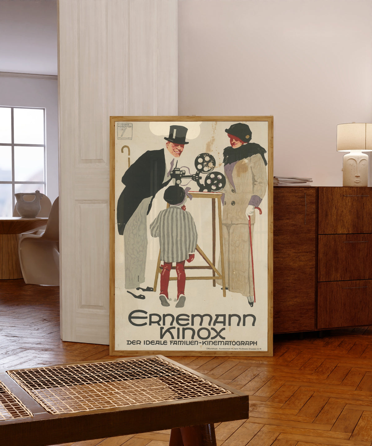 Framed art print of Ernemann Kinox: Der ideale Familien-Kinematograph by Ludwig Hohlwein, in a light wood frame leaning in a warm room with a parquet floor