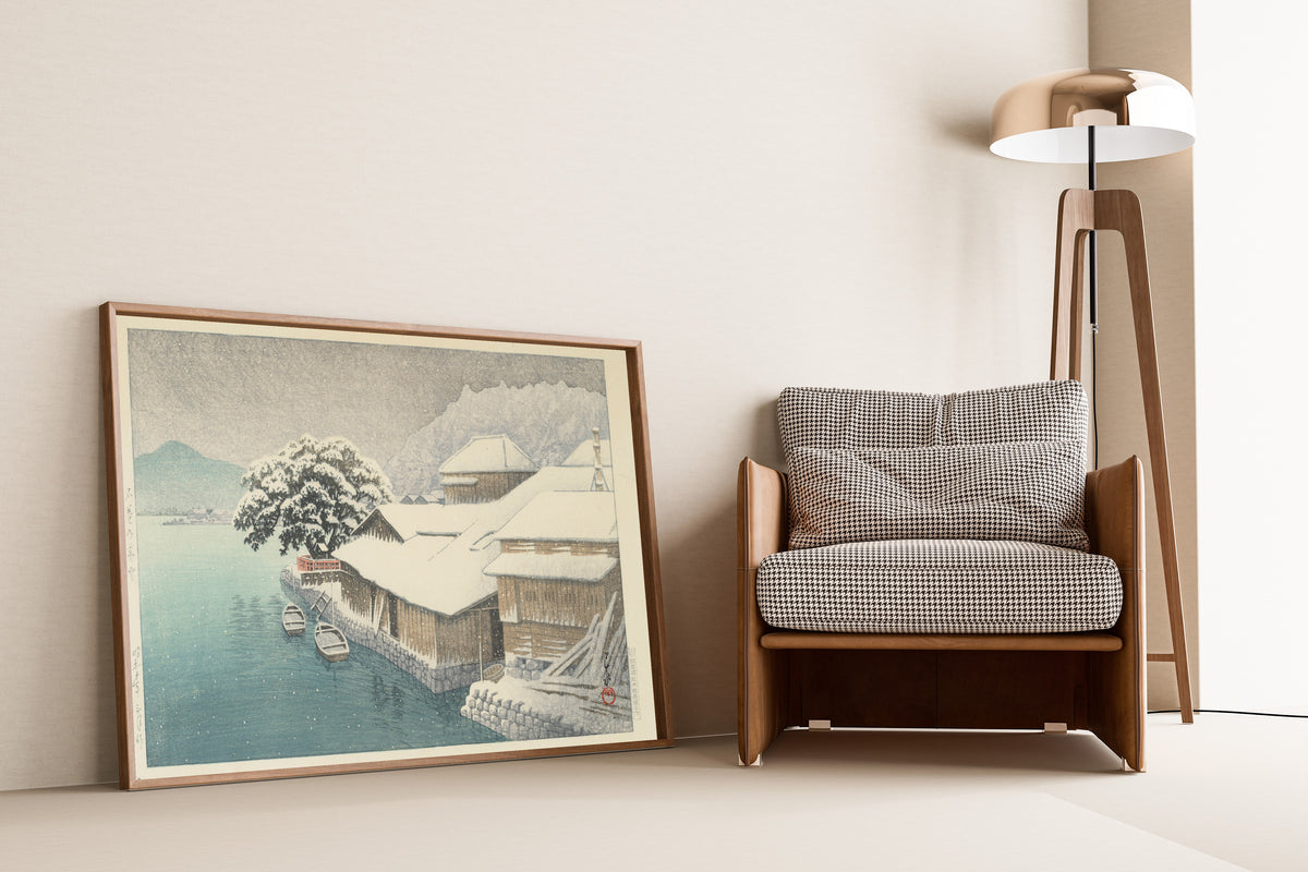 Framed art print of Evening Snow at Ishinomaki by Kawase Hasui, leaning against a wall beside an armchair and a floor lamp