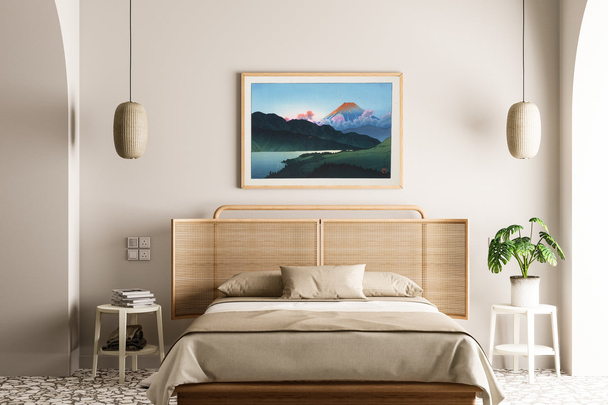Framed art print of Evening of Mount Fuji from Lake Ashino-ko by Kawase Hasui, above a cane-headboard bed
