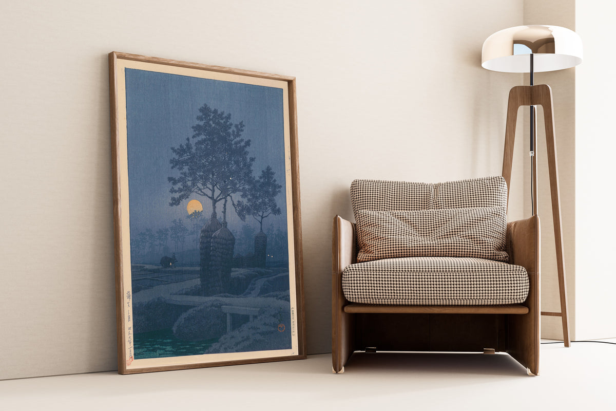 Framed art print of Full Moon at Gamo by Kawase Hasui, leaning against a wall beside an armchair and a floor lamp