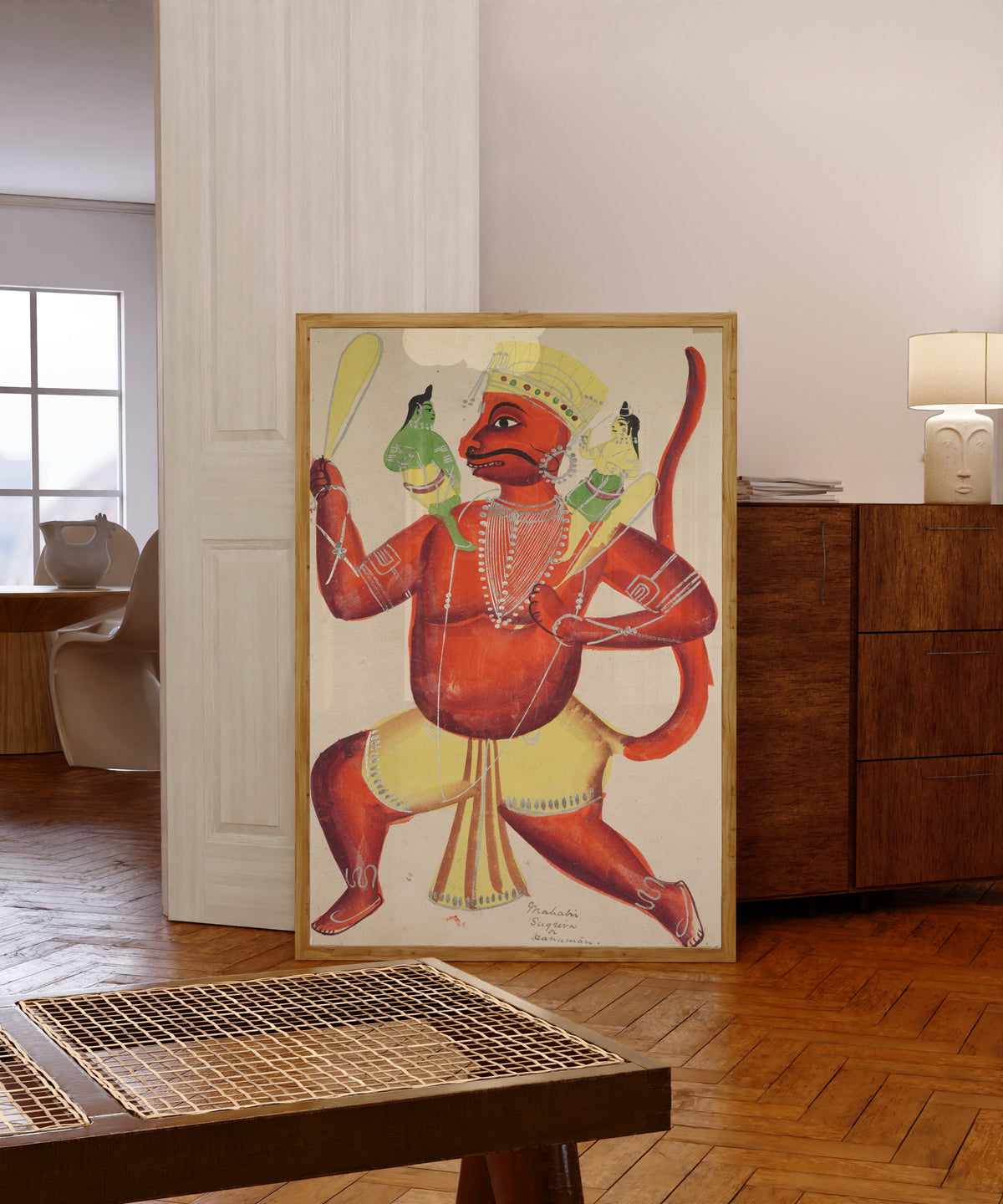 Framed art print of Hanuman with Rama and Lakshmana on his shoulders… by Kalighat painter, in a light wood frame leaning in a warm room with a parquet floor