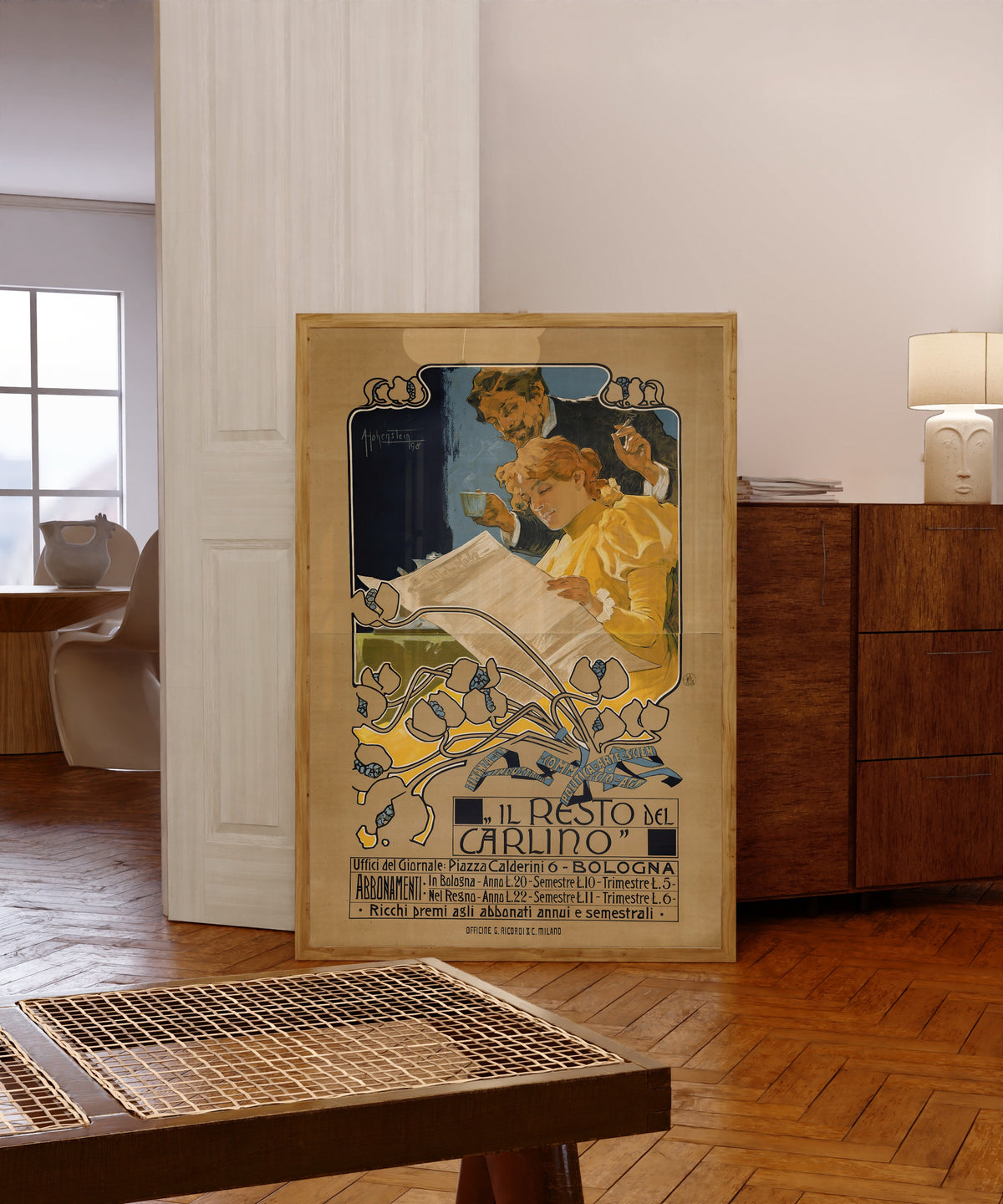Framed art print of Il Resto del Carlino, Bologna by Adolfo Hohenstein, in a light wood frame leaning in a warm room with a parquet floor