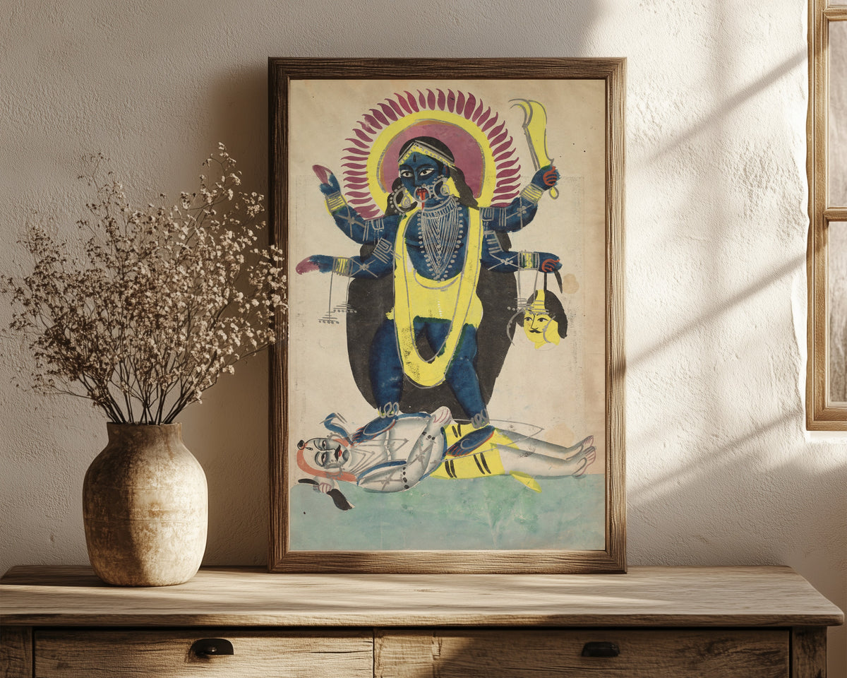 Framed art print of Kali Dancing on Shiva by Kalighat painter, in a dark wood frame leaning on a rustic console beside dried flowers