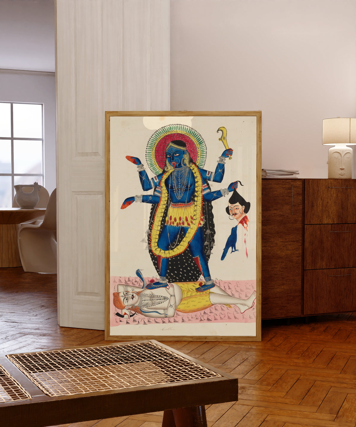 Framed art print of Kali Trampling Upon Shiva by Kalighat painter, in a light wood frame leaning in a warm room with a parquet floor