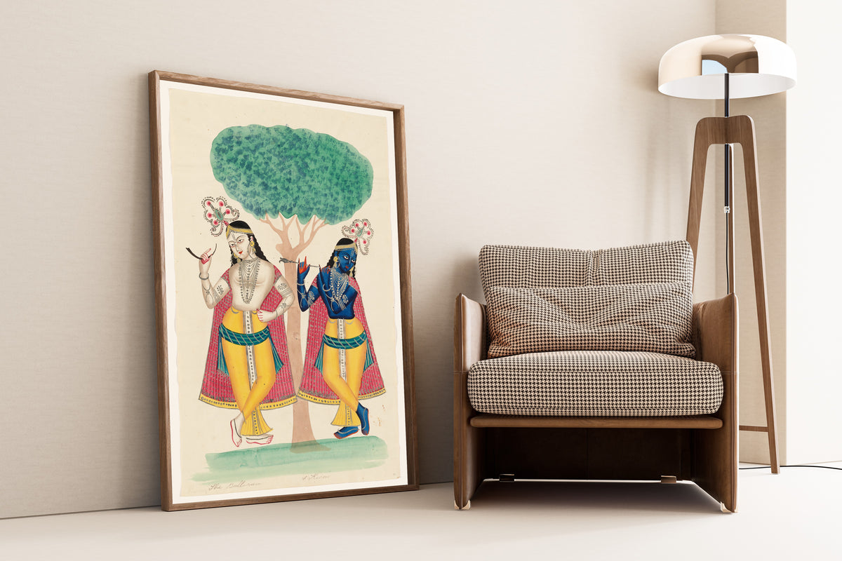 Framed art print of Krishna and Balarama underneath a Tree by Kalighat painter, leaning against a wall beside an armchair and a floor lamp