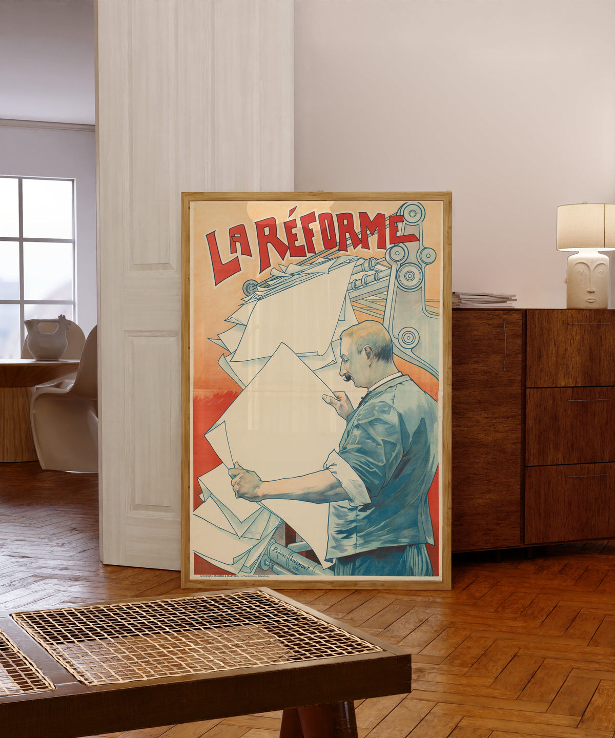 Framed art print of La Réforme by Henri Privat-Livemont, in a light wood frame leaning in a warm room with a parquet floor