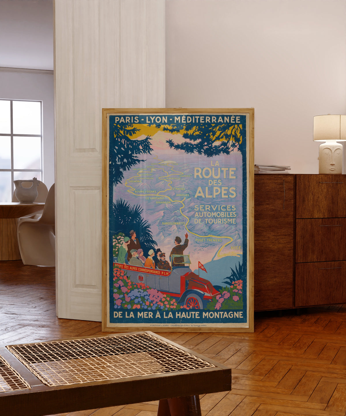 Framed art print of La Route des Alpes by Roger Broders, in a light wood frame leaning in a warm room with a parquet floor