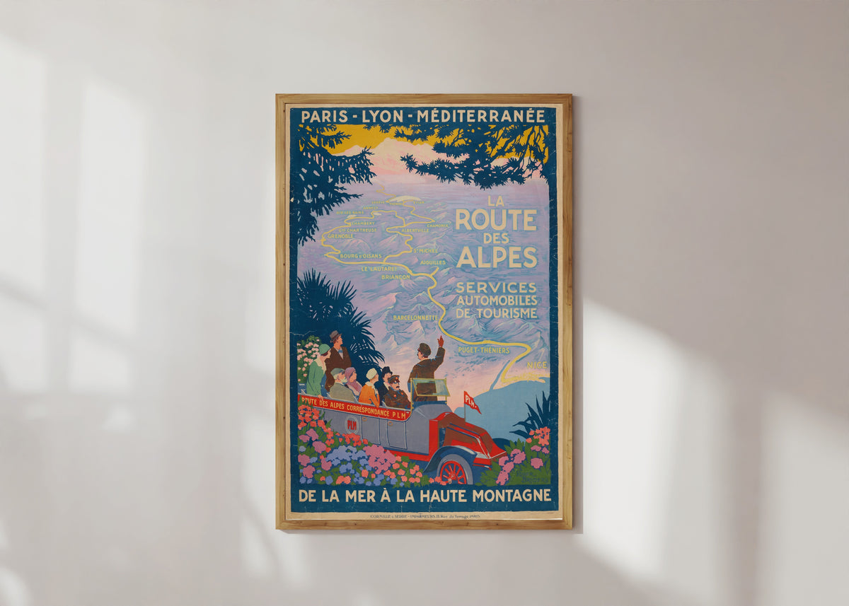 Framed art print of La Route des Alpes by Roger Broders, in a light wood frame on a sunlit white wall