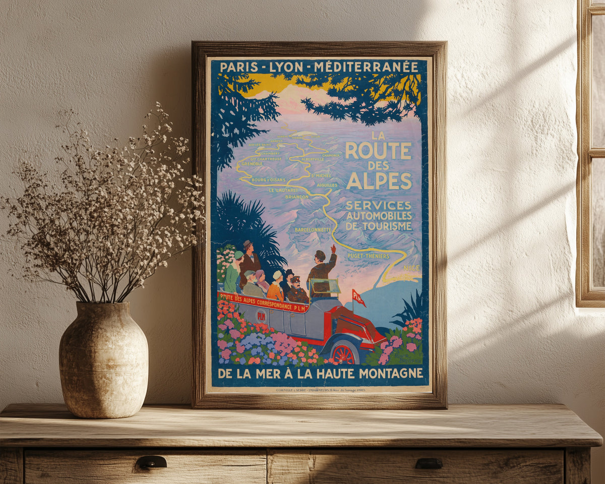 Framed art print of La Route des Alpes by Roger Broders, in a dark wood frame leaning on a rustic console beside dried flowers