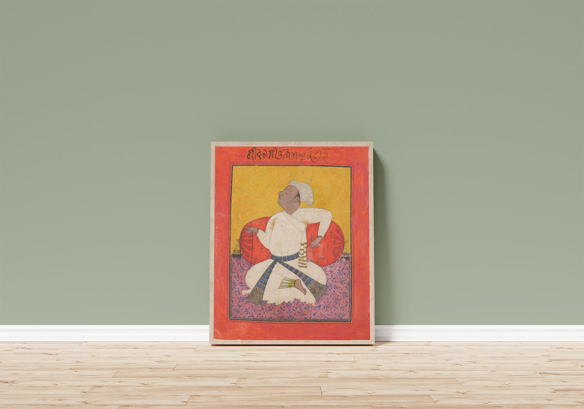 Framed art print of Maharaja Sital Dev in Devotion by Basohli school, in a light wood frame leaning on a sage green wall above a wooden floor