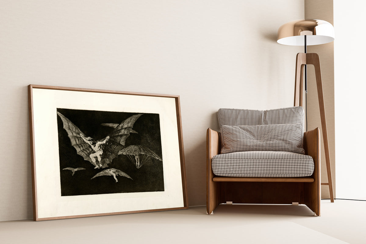 Framed art print of Modo de volar (A Way of Flying), from Los Disparates by Francisco de Goya, leaning against a wall beside an armchair and a floor lamp