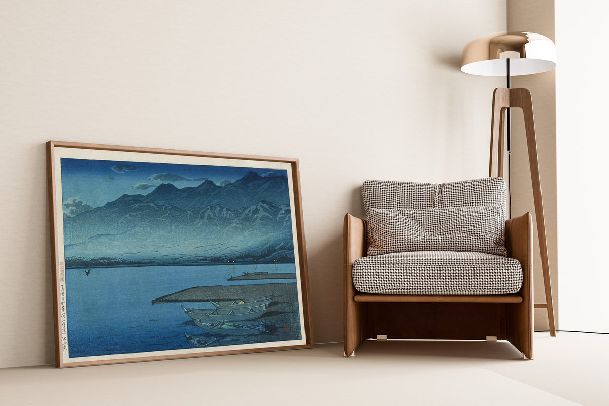 Framed art print of Moonlight over Lake Kamo, Sado by Kawase Hasui, leaning against a wall beside an armchair and a floor lamp
