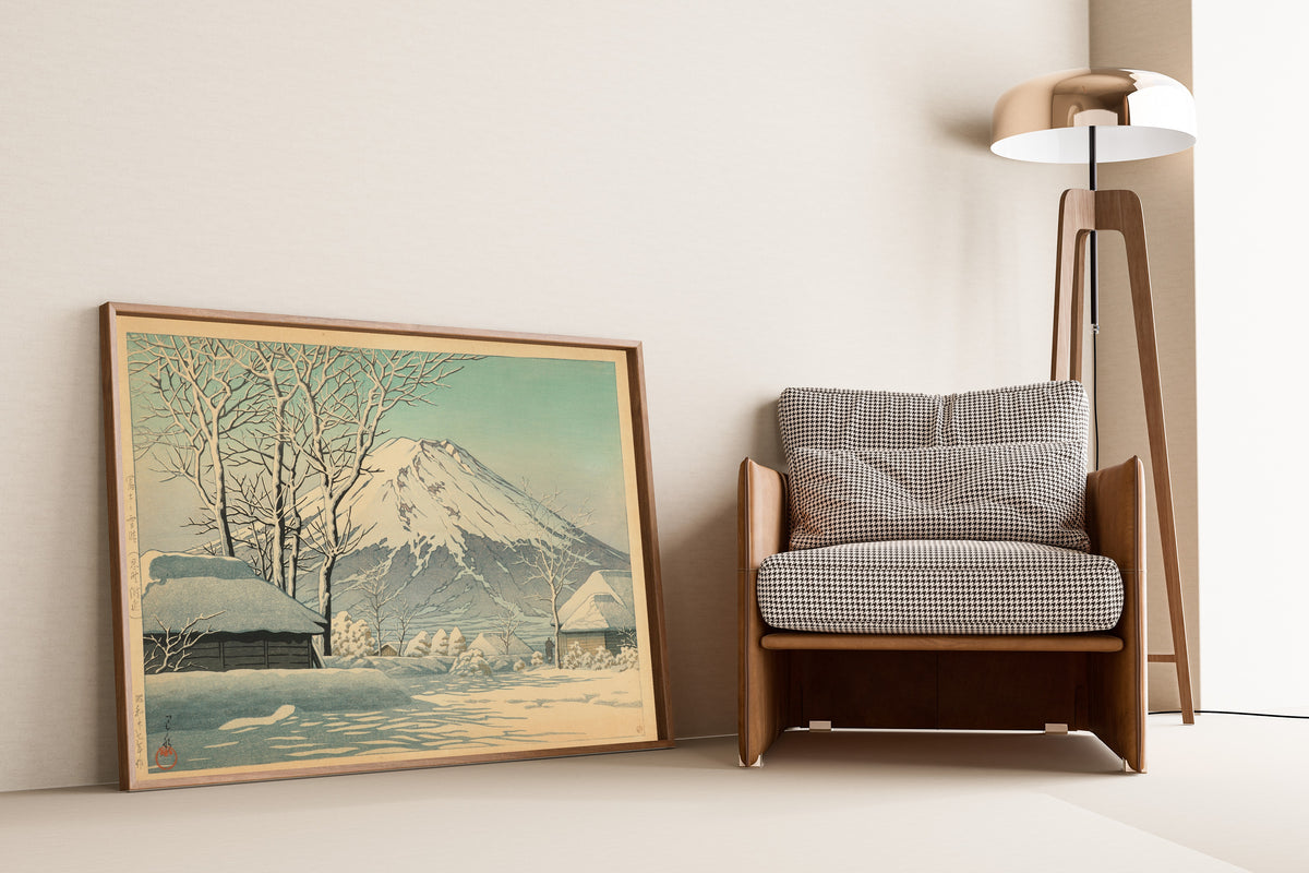 Framed art print of Mount Fuji, Clearing after a Snowfall, Oshino by Kawase Hasui, leaning against a wall beside an armchair and a floor lamp