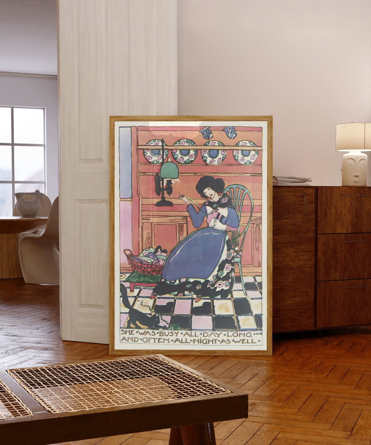 Framed art print of Mummy's Bedtime Story Book: She Was Busy All Day Long… by Jessie M. King, in a light wood frame leaning in a warm room with a parquet floor