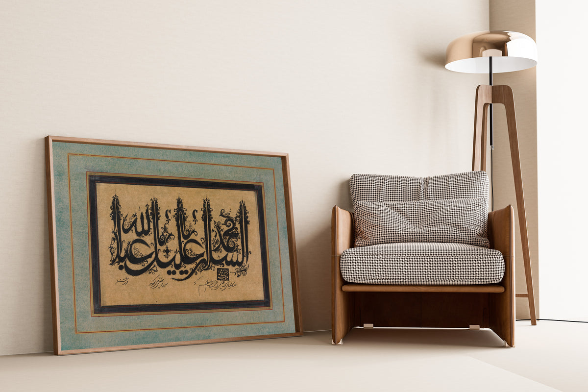 Framed art print of Page of Calligraphy in Ornamental Style by Malik Muhammad Qazvini, leaning against a wall beside an armchair and a floor lamp