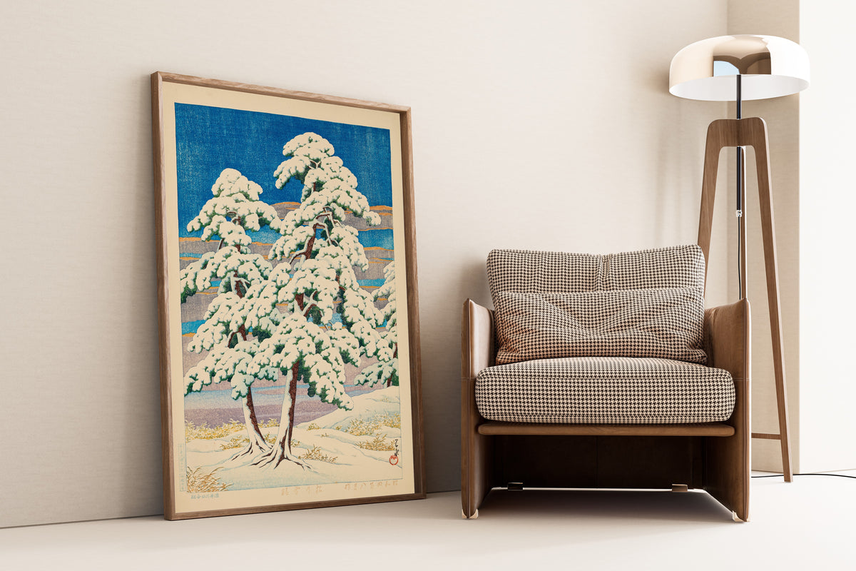 Framed art print of Pines in Clear Weather After Snow by Kawase Hasui, leaning against a wall beside an armchair and a floor lamp