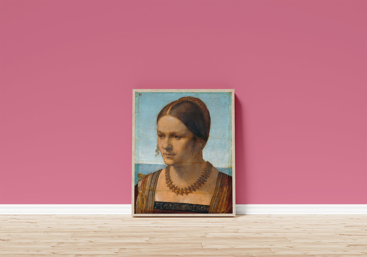 Framed art print of Portrait of a Young Venetian Woman by Albrecht Dürer, in a light wood frame leaning on a pink wall above a wooden floor