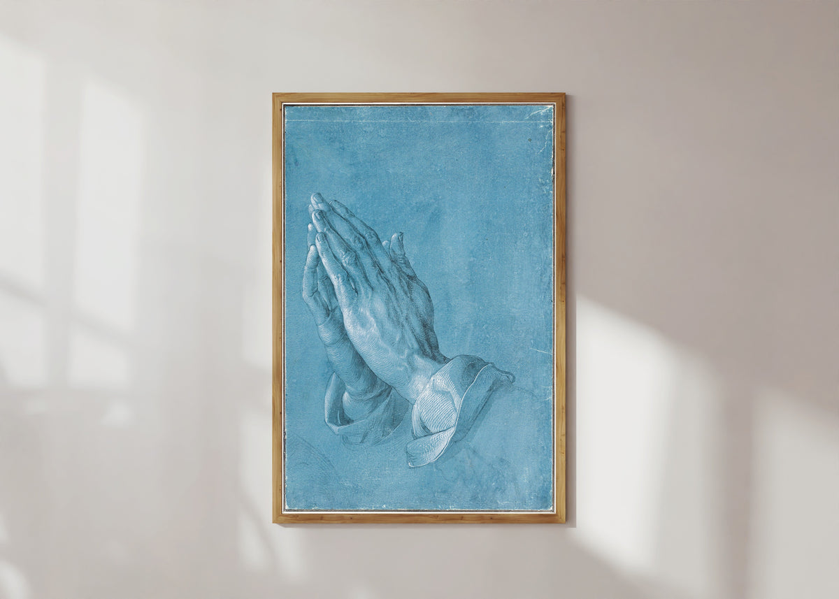 Framed art print of Praying Hands by Albrecht Dürer, in a light wood frame on a sunlit white wall