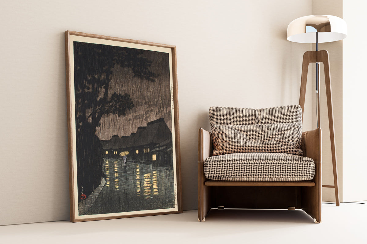 Framed art print of Rain at Maekawa, Sagami Province by Kawase Hasui, leaning against a wall beside an armchair and a floor lamp