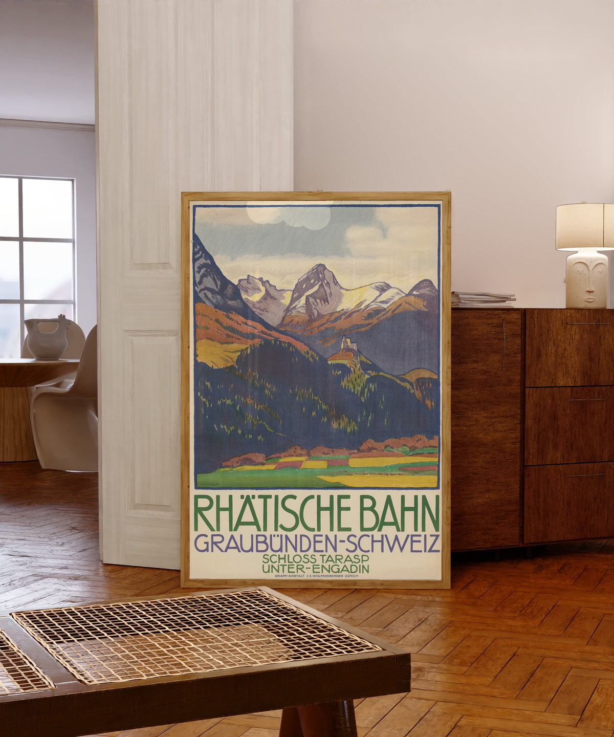 Framed art print of Rhätische Bahn: Graubünden-Schweiz, Schloss Tarasp… by Emil Cardinaux, in a light wood frame leaning in a warm room with a parquet floor