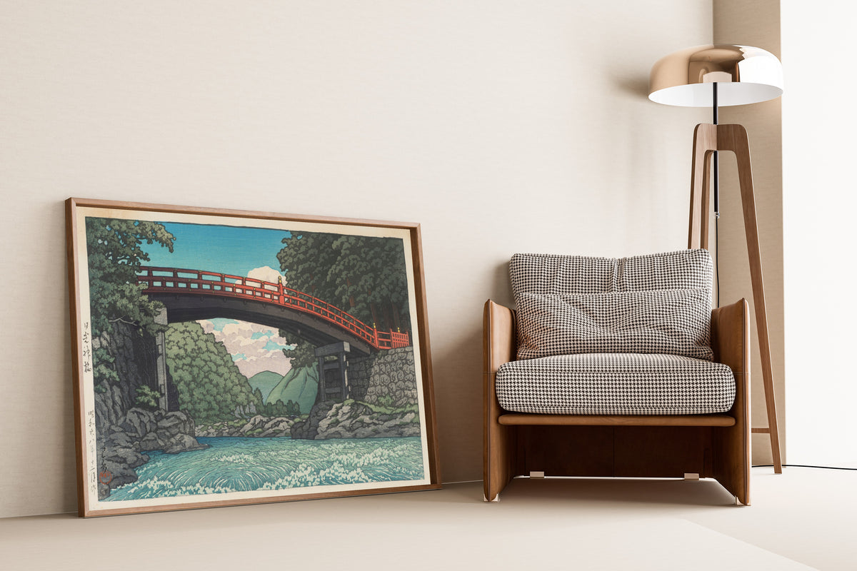 Framed art print of Sacred Bridge, Nikko by Kawase Hasui, leaning against a wall beside an armchair and a floor lamp