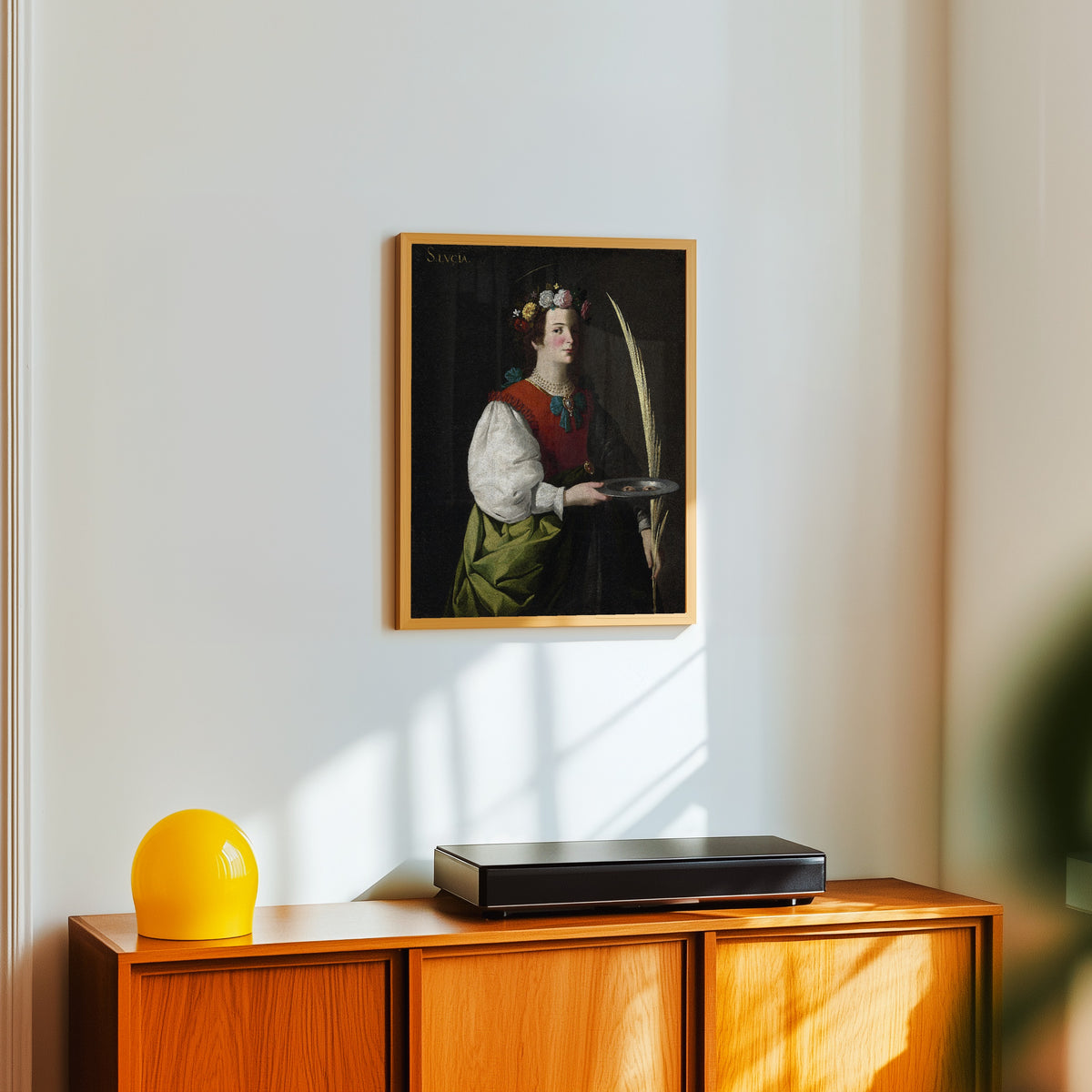 Framed art print of Saint Lucy by Francisco de Zurbarán, in a light wood frame on a white wall above an orange sideboard