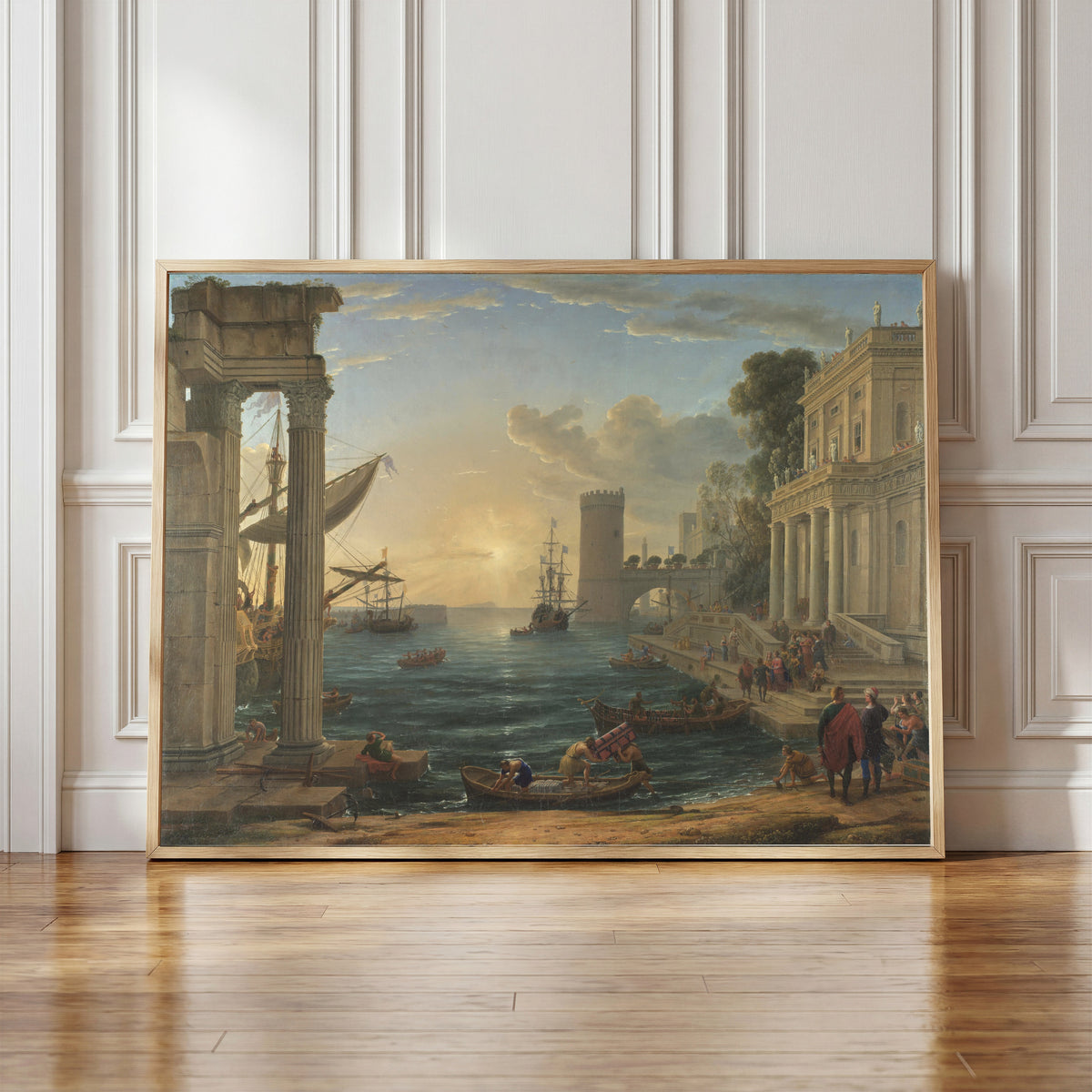 Framed art print of Seaport with the Embarkation of the Queen of Sheba by Claude Lorrain, in a light wood frame leaning against a panelled white wall
