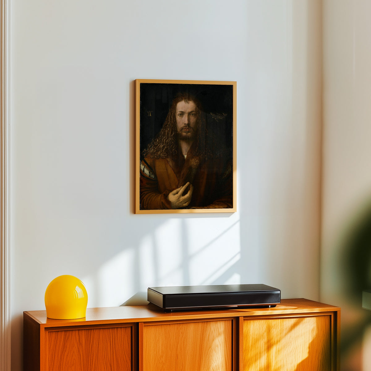 Framed art print of Self-Portrait in a Fur-Trimmed Coat by Albrecht Dürer, in a light wood frame on a white wall above an orange sideboard