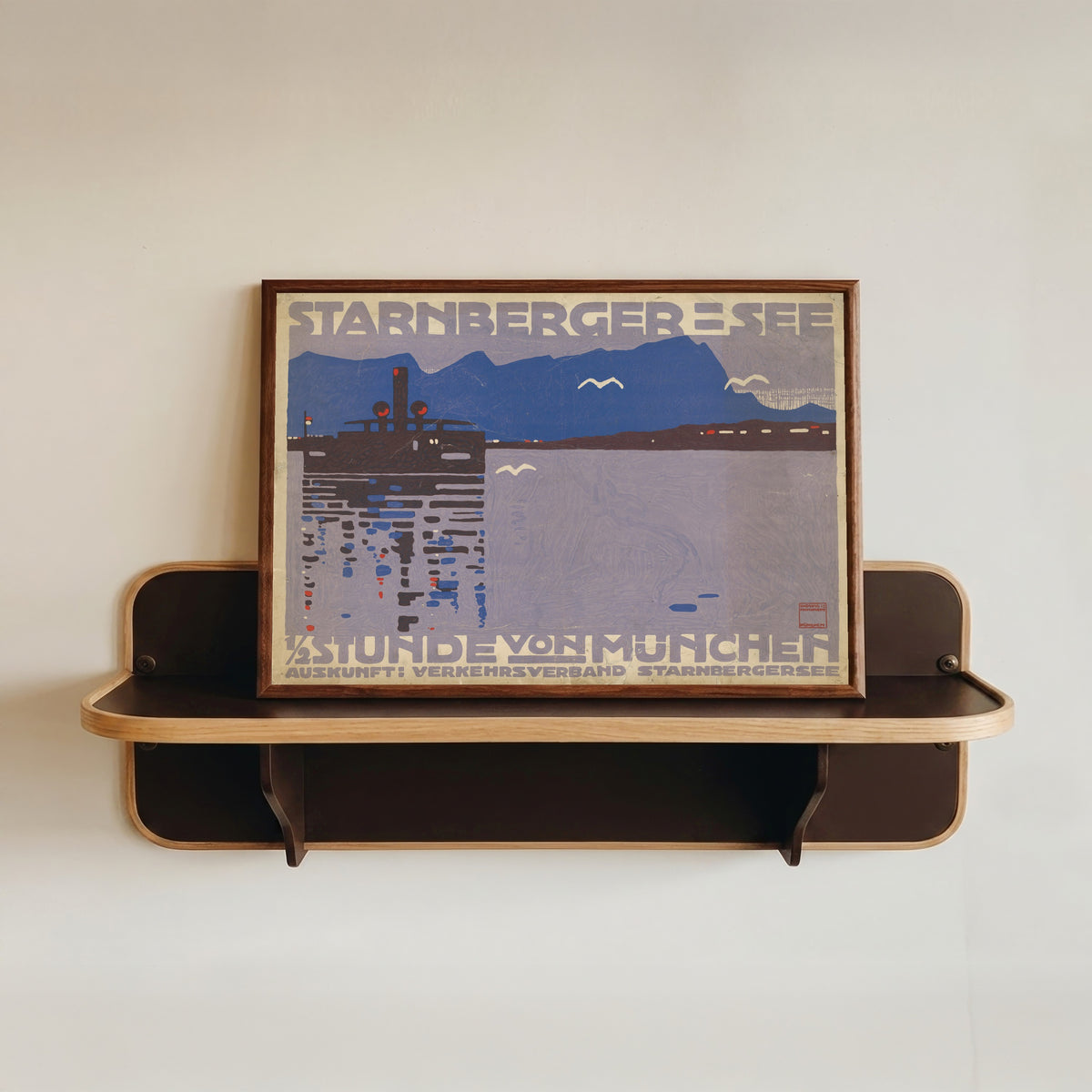 Framed art print of Starnberger See: ½ Stunde von München by Ludwig Hohlwein, in a dark wood frame resting on a wall shelf