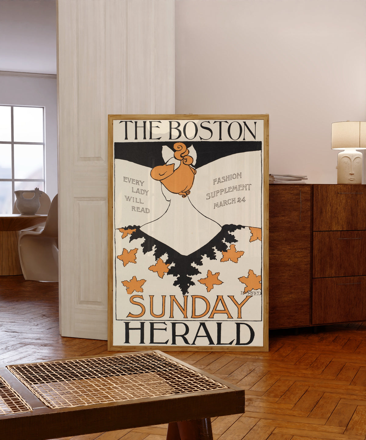 Framed art print of The Boston Sunday Herald, Every Lady Will Read, Fashion… by Ethel Reed, in a light wood frame leaning in a warm room with a parquet floor