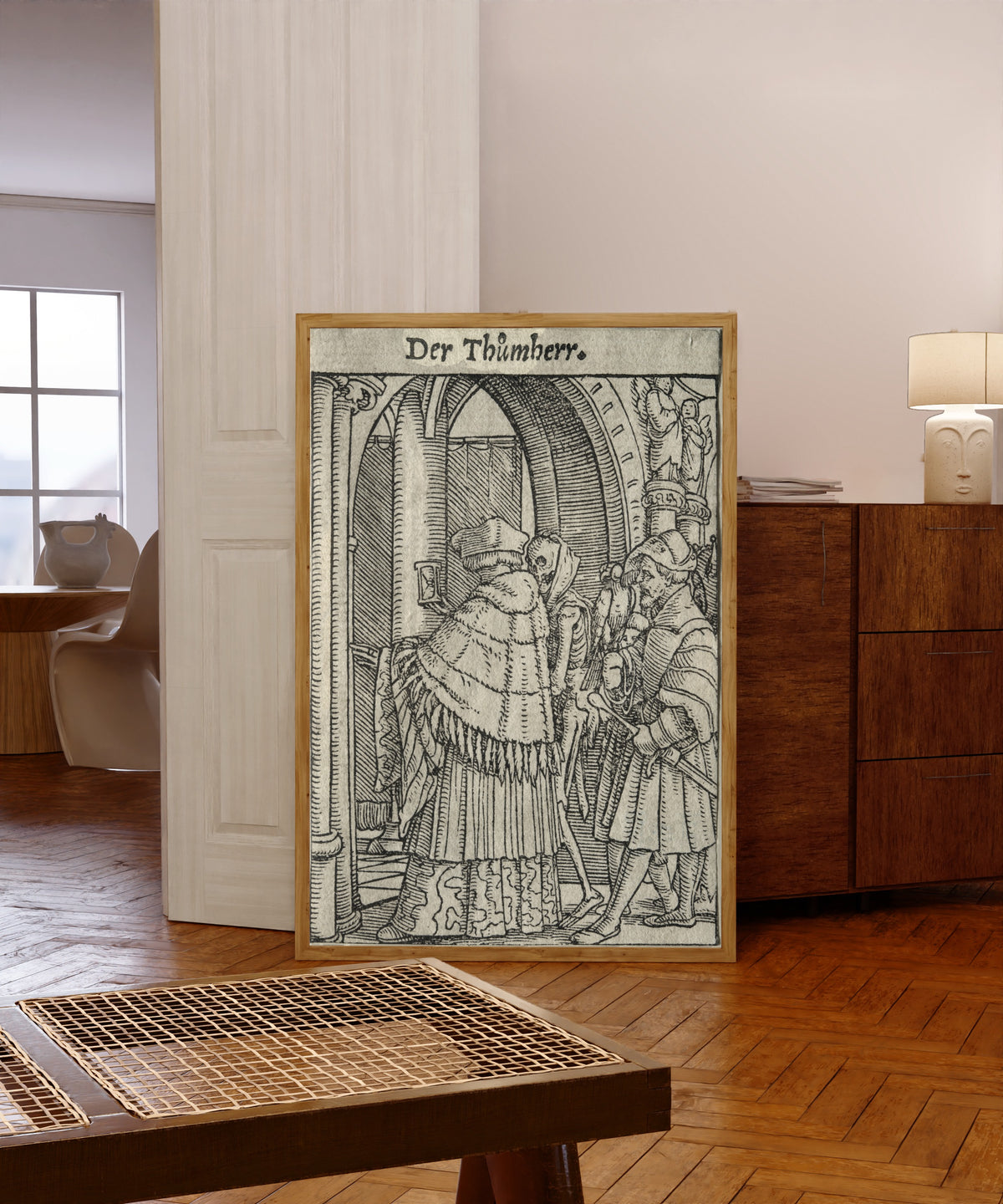 Framed art print of The Dance of Death: Der Thůmherr (The Canon) by Hans Holbein the Younger, in a light wood frame leaning in a warm room with a parquet floor