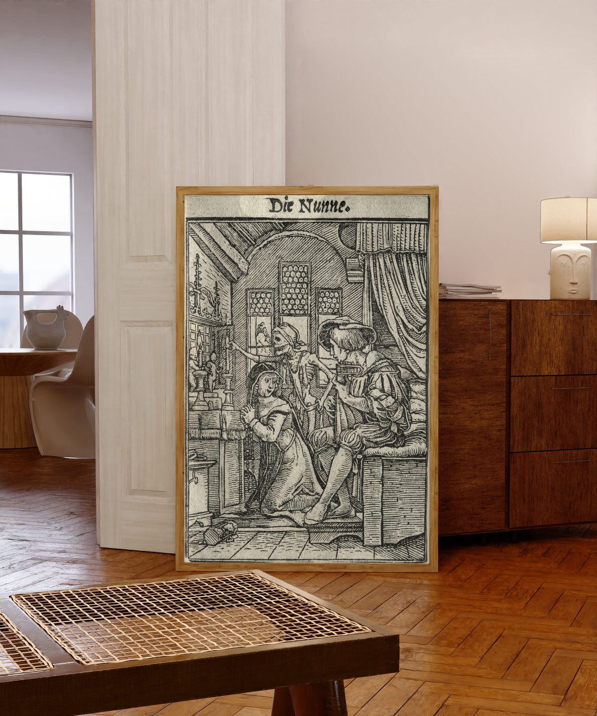 Framed art print of The Dance of Death: Die Nunne (The Nun) by Hans Holbein the Younger, in a light wood frame leaning in a warm room with a parquet floor