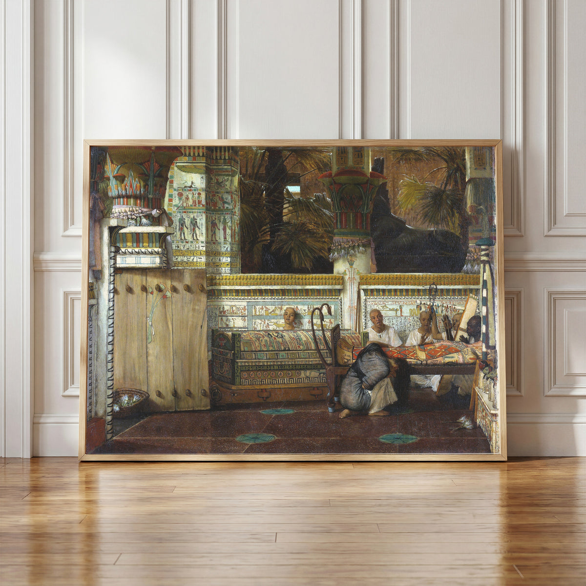 Framed art print of The Egyptian Widow by Lawrence Alma-Tadema, in a light wood frame leaning against a panelled white wall