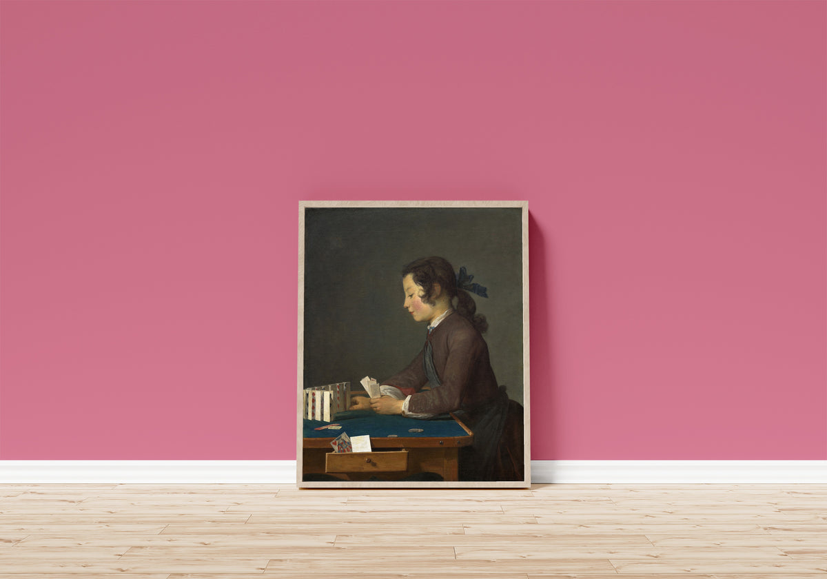 Framed art print of The House of Cards by Jean-Siméon Chardin, in a light wood frame leaning on a pink wall above a wooden floor
