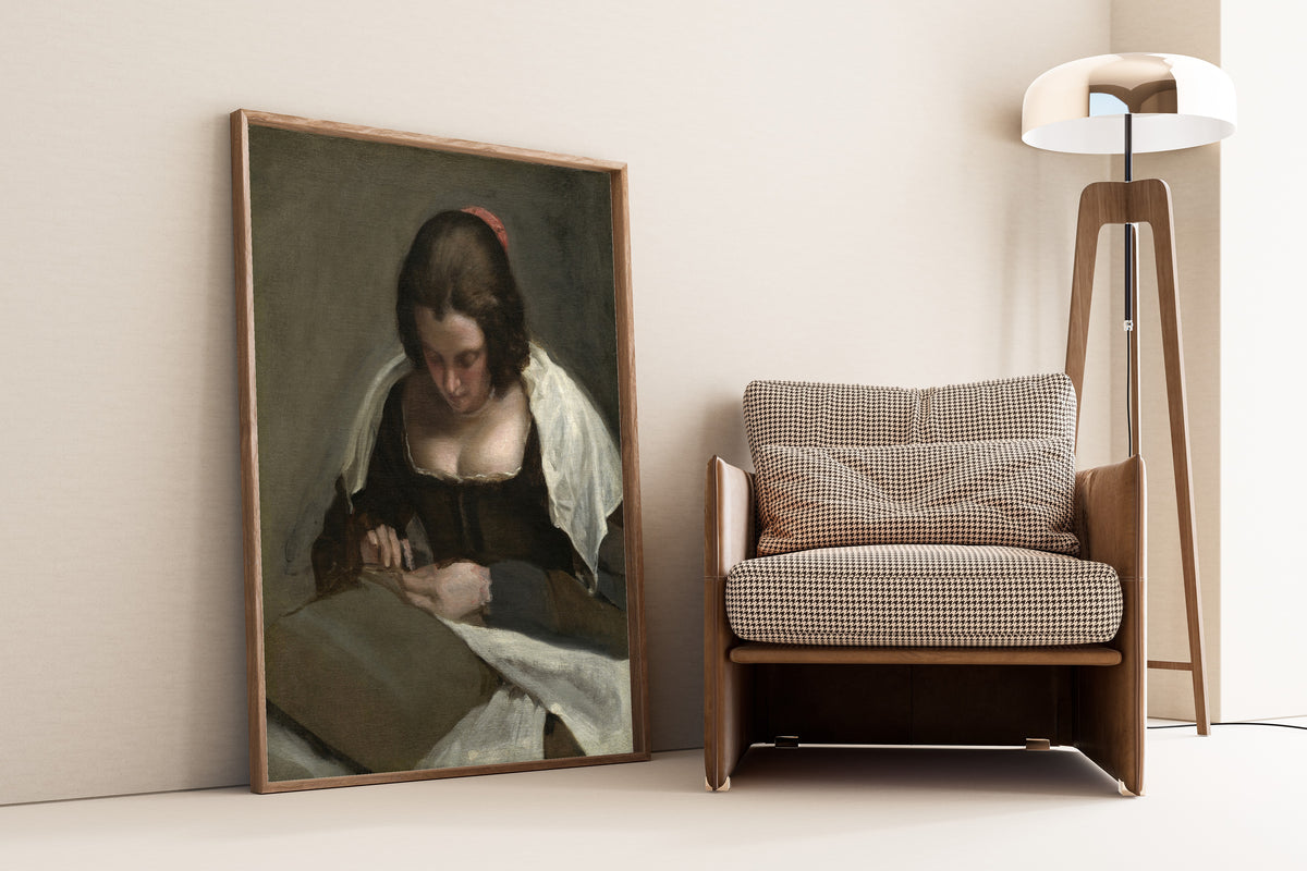 Framed art print of The Needlewoman by Diego Velázquez, leaning against a wall beside an armchair and a floor lamp