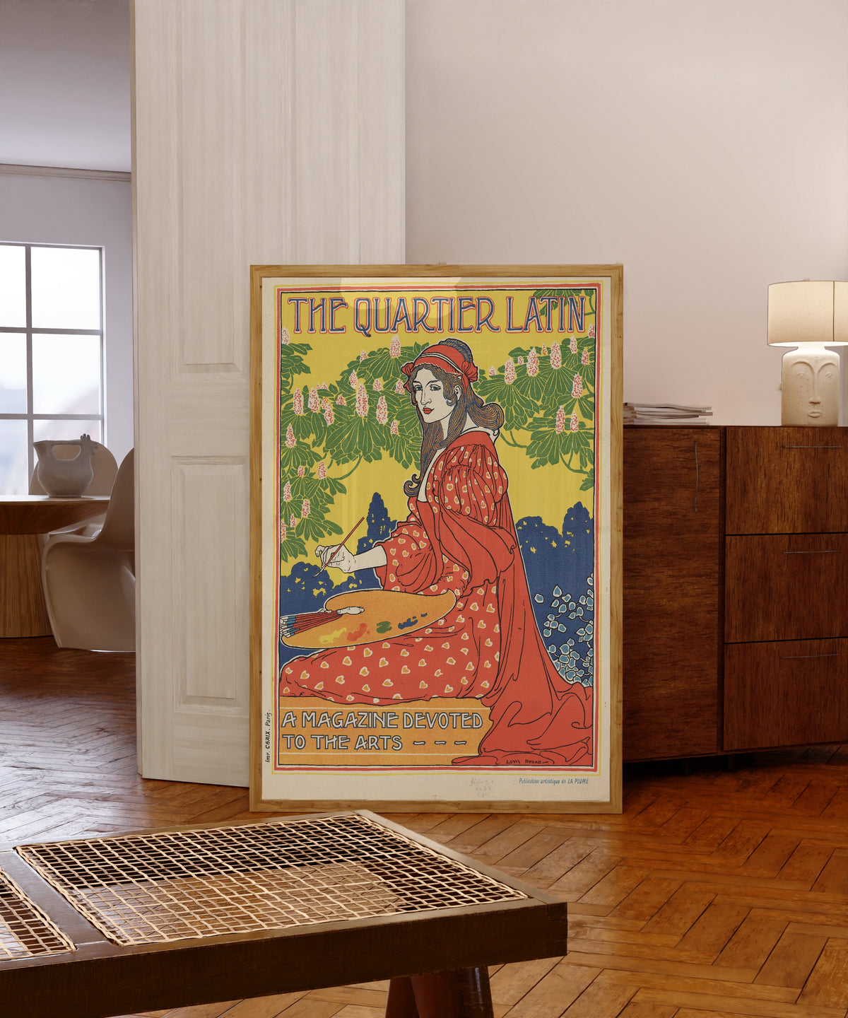 Framed art print of The Quartier Latin, A Magazine Devoted to the Arts by Louis Rhead, in a light wood frame leaning in a warm room with a parquet floor