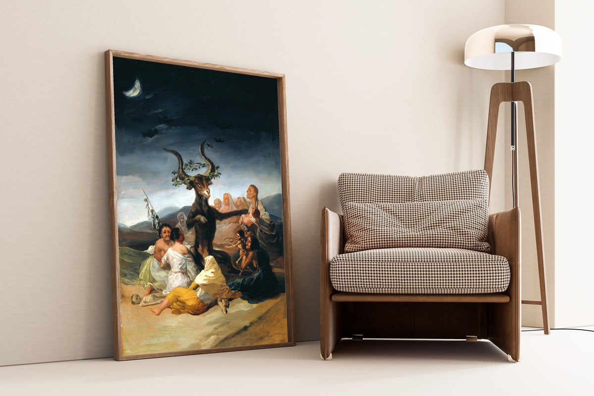 Framed art print of The Sabbath of Witches by Francisco de Goya, leaning against a wall beside an armchair and a floor lamp