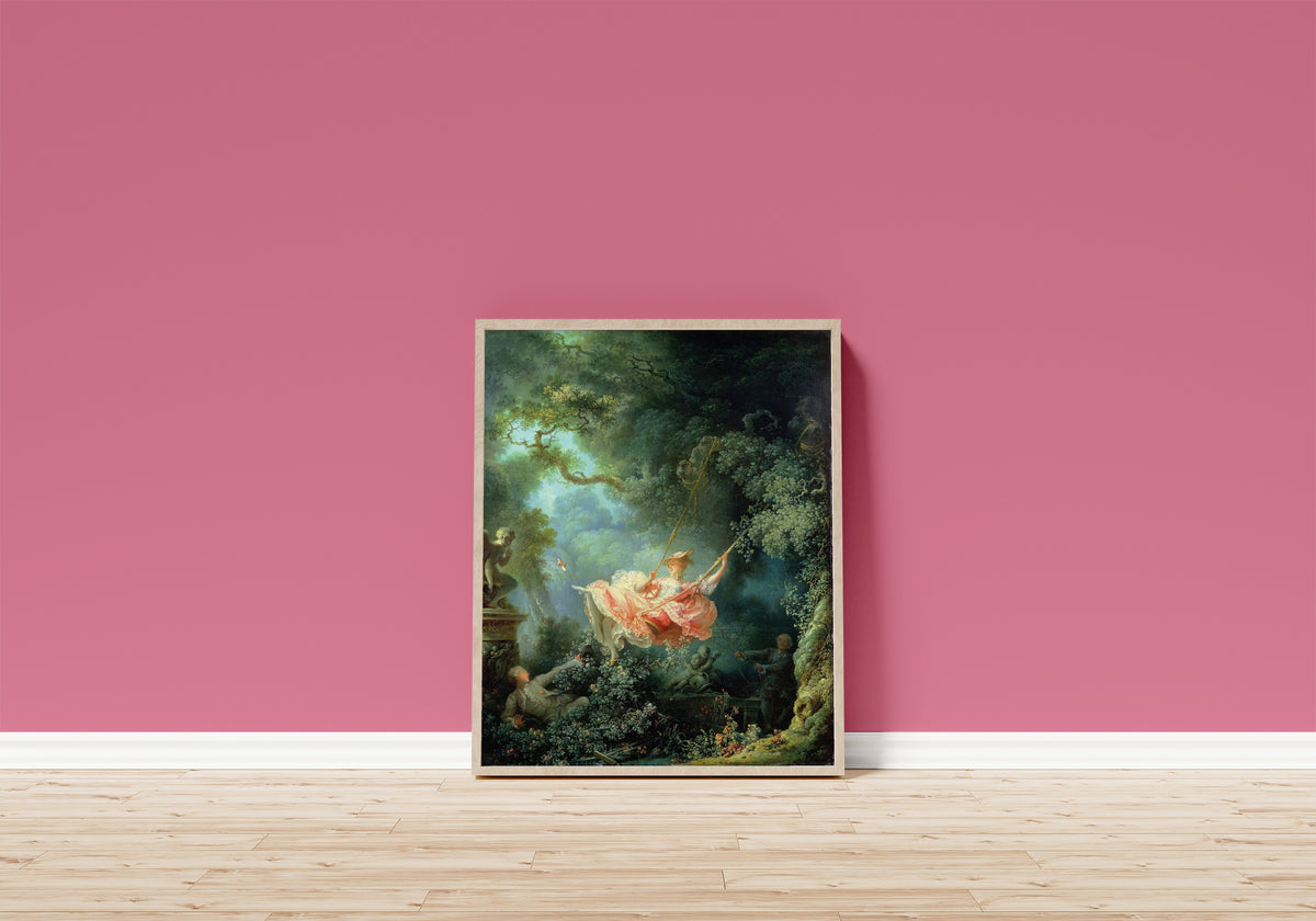Framed art print of The Swing by Jean-Honoré Fragonard, in a light wood frame leaning on a pink wall above a wooden floor