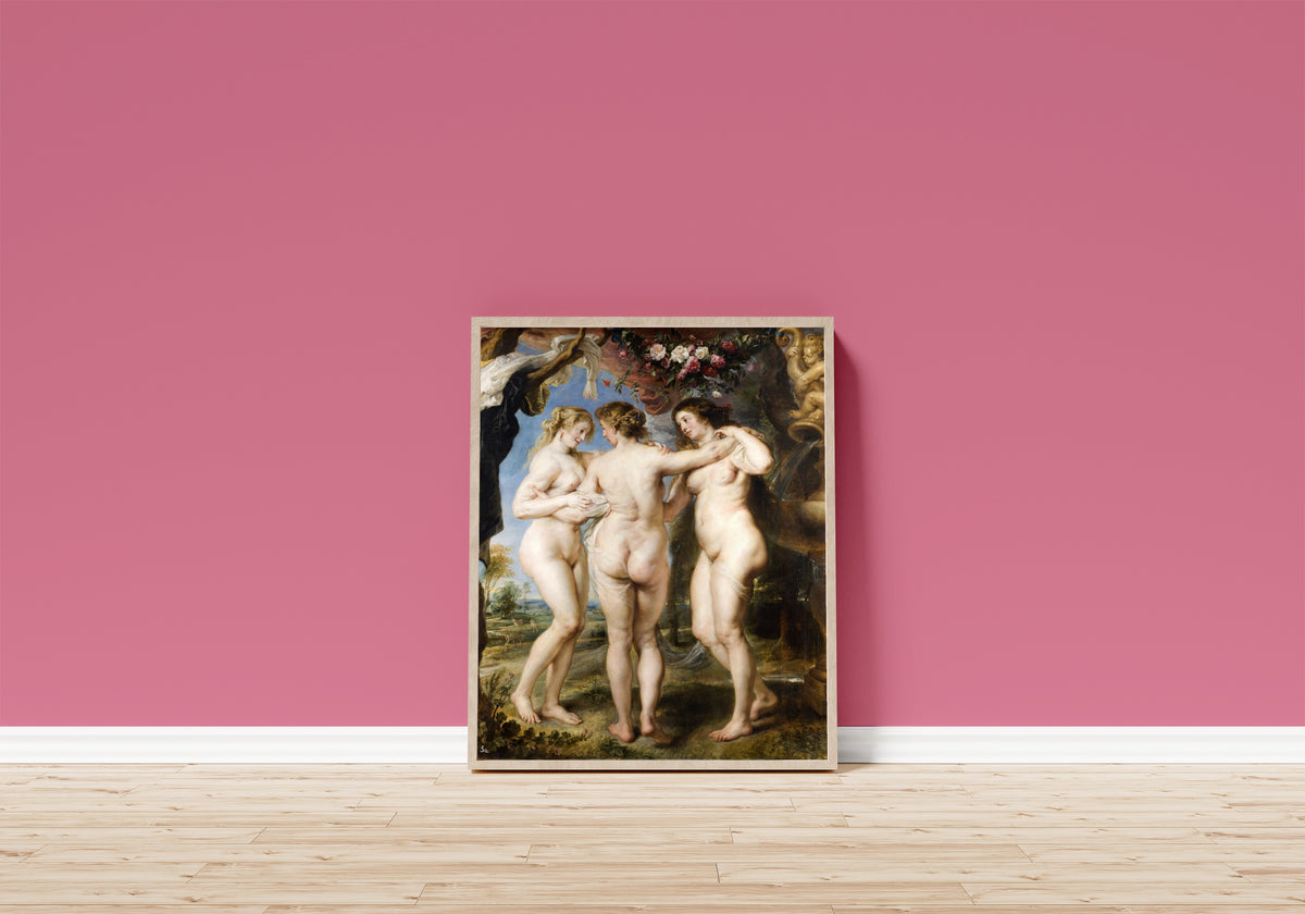 Framed art print of The Three Graces by Peter Paul Rubens, in a light wood frame leaning on a pink wall above a wooden floor